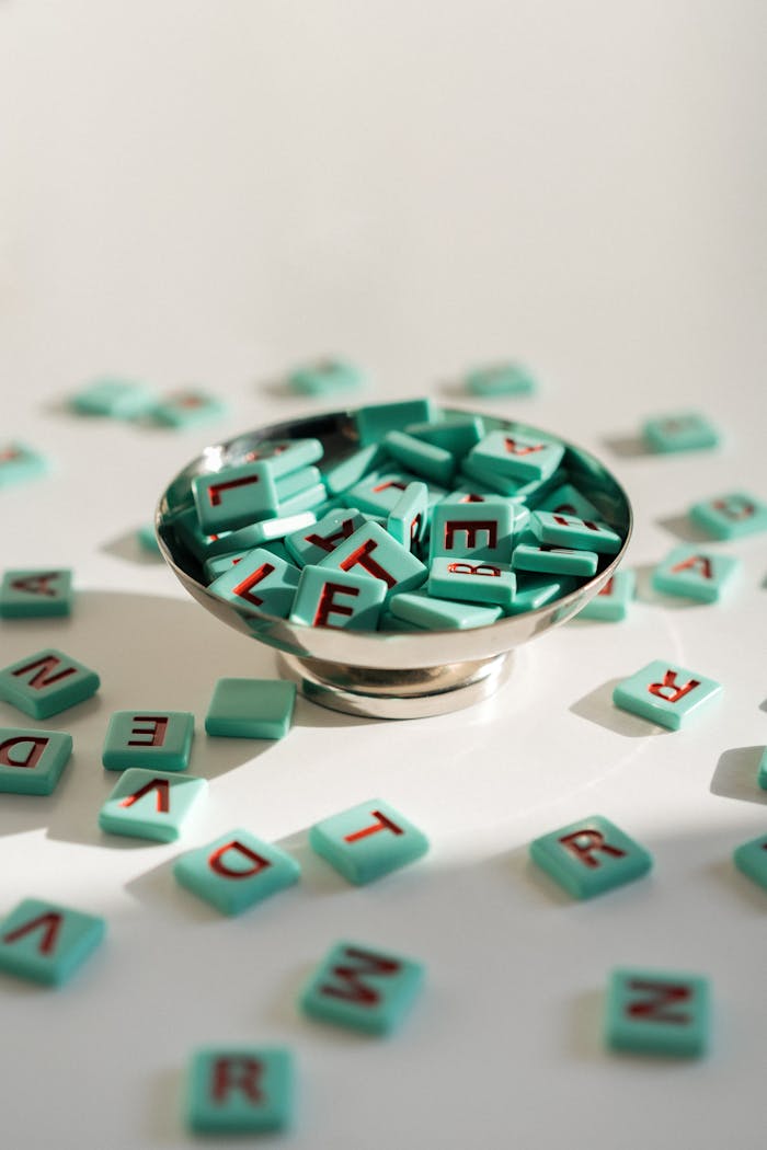 menu-13 Bright photo of turquoise letter tiles scattered around a silver bowl on a light surface.